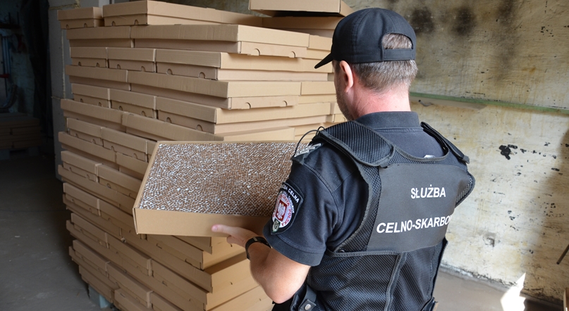 Funkcjonariusz KAS trzyma w dłoniach karton z nielegalnymi papierosami. W tle cała paleta podobnych kartonów.