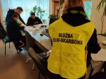 Na zdjęciu grupa osób siedzi przy stole. Obok stoi funkcjonariusz Służby Celno-Skarbowej.