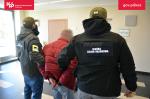 dwaj funkcjonariusze SCS prowadzą skutego w kajdanki mężczyznę w czerwonej kurtce