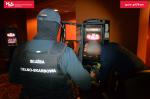 Funkcjonariusz Służby Celno-Skarbowej stoi przed automatem do nielegalnego urządzania gier hazardowych, inny mężczyzna wyjmuje kasetkę z automatu