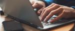 Na zdjęciu widoczne są dłonie na laptopie i leżący obok telefon komórkowy